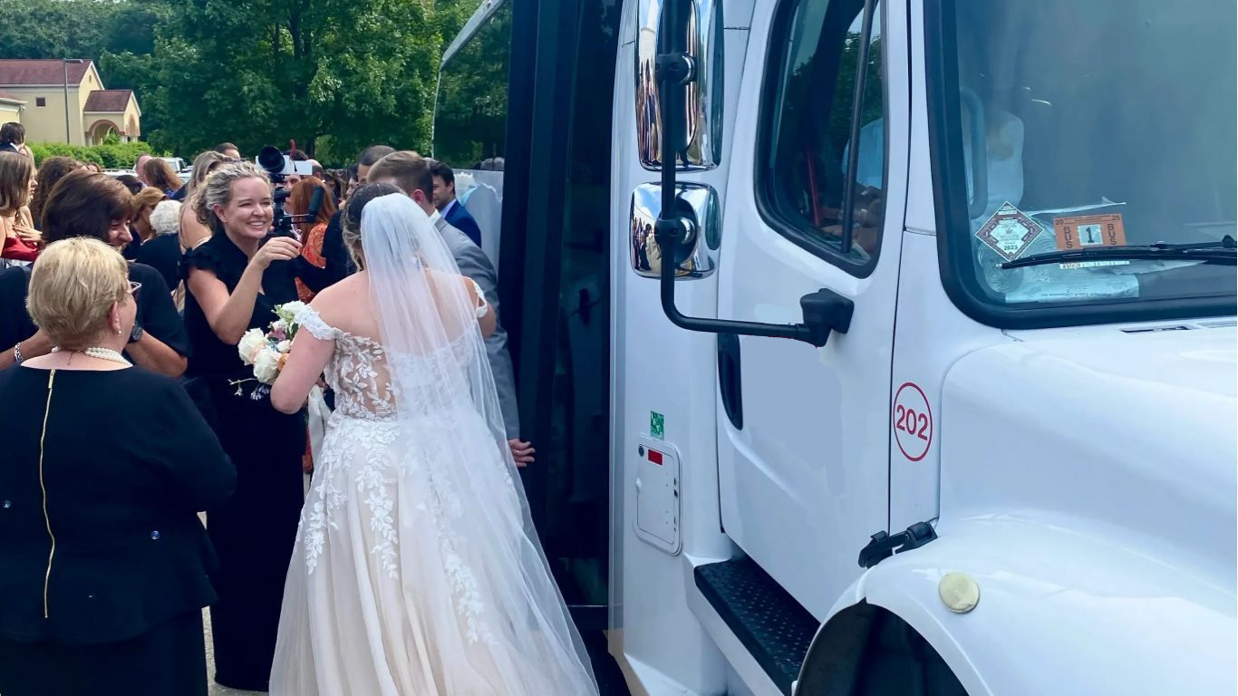 A bride in a white wedding gown and veil boards a white bus, while guests stand nearby on a paved area under a sunny sky.