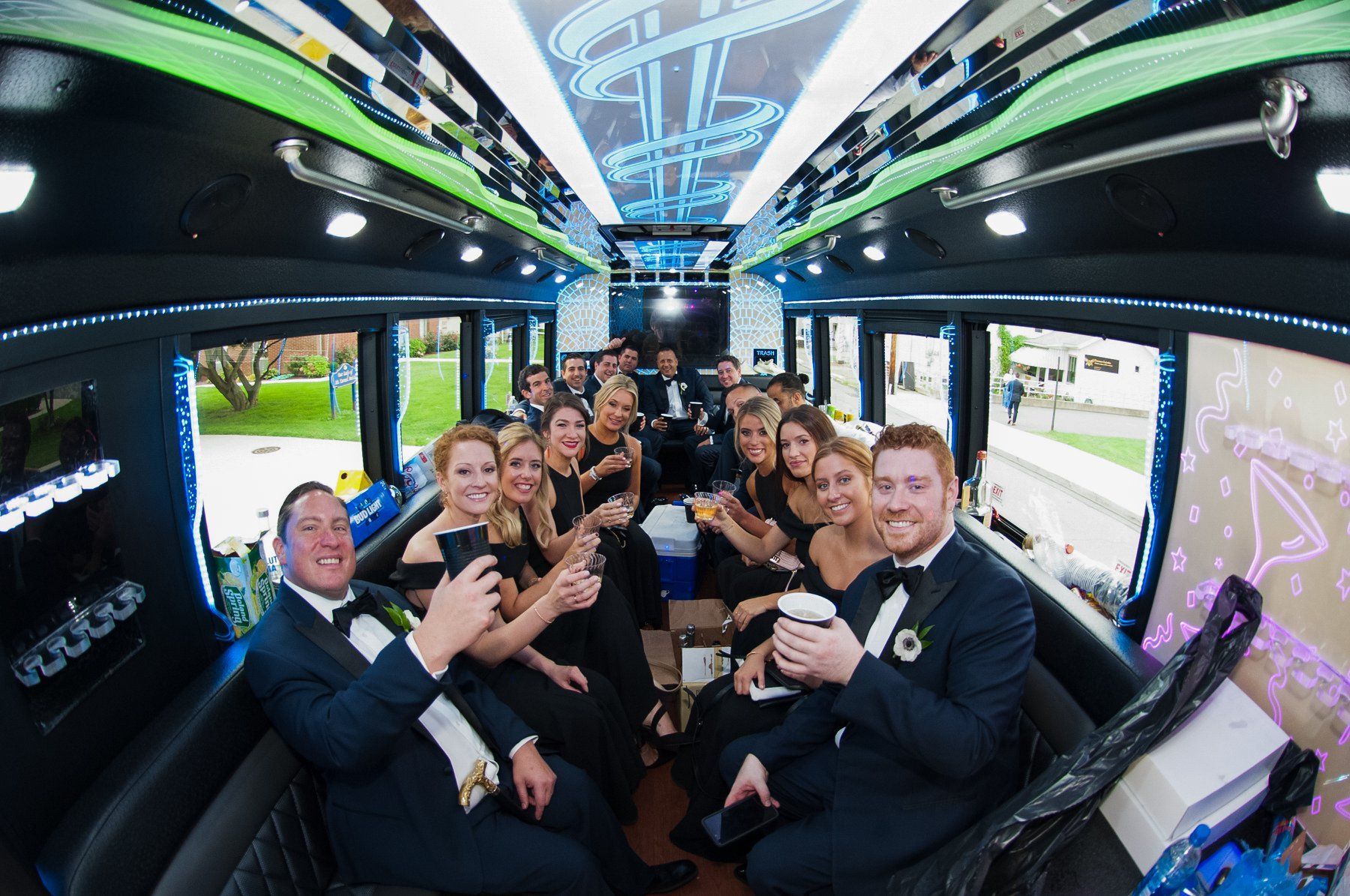 A group in formal wedding attire toast with drinks while sitting inside a brightly lit party bus.