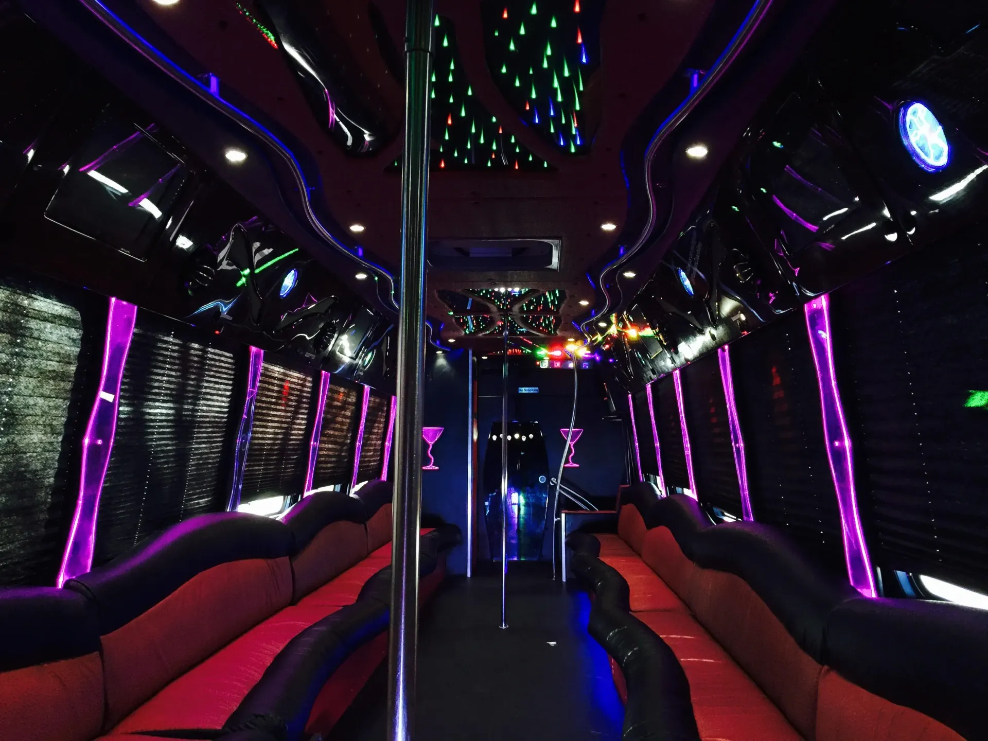 The interior of a party bus featuring long red-and-black leather bench seating, a central dance pole, and colorful lighting.