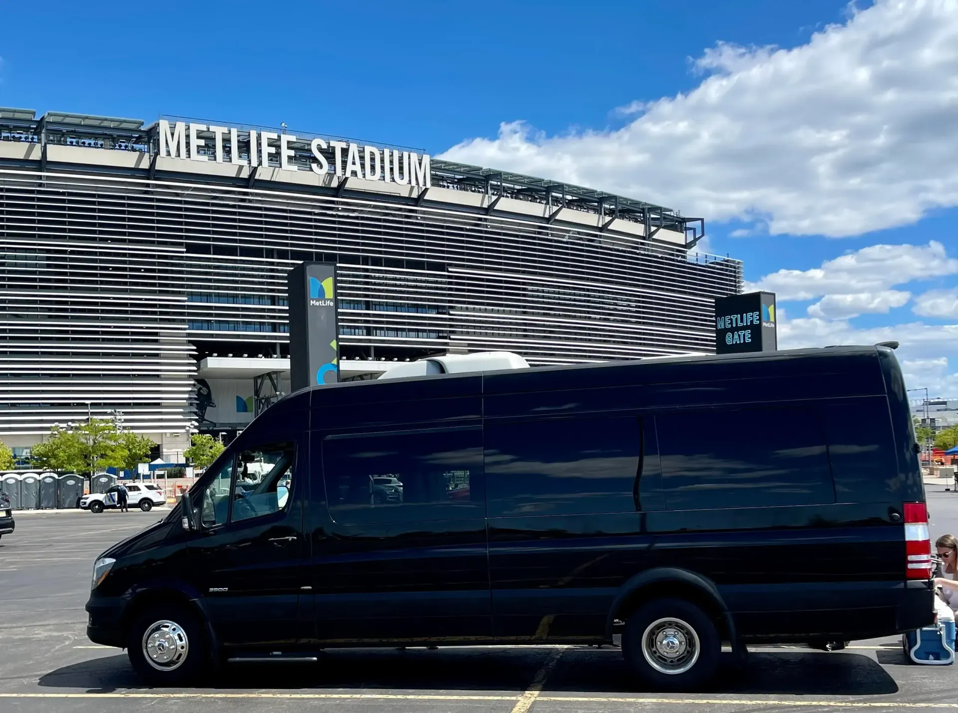 A black passenger van parked in a paved lot in front of the MetLife Stadium under a blue sky.