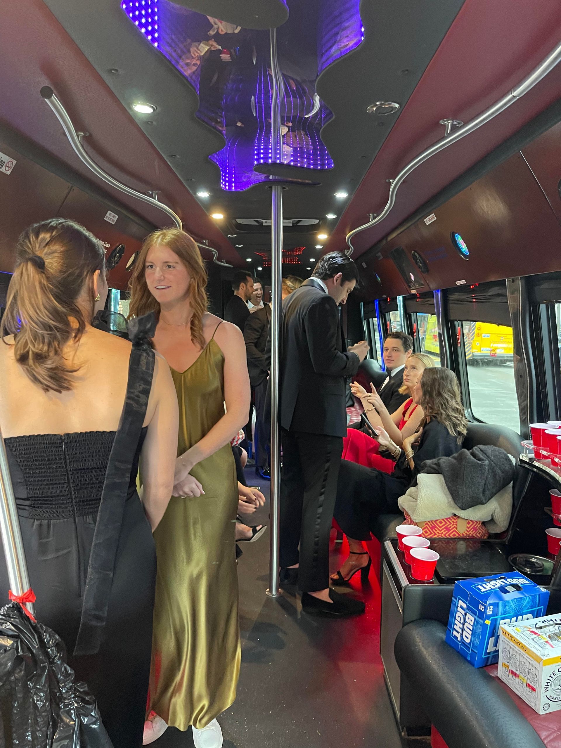People in formal wear stand and sit inside a dimly lit party bus with red accents and LED ceiling lights.