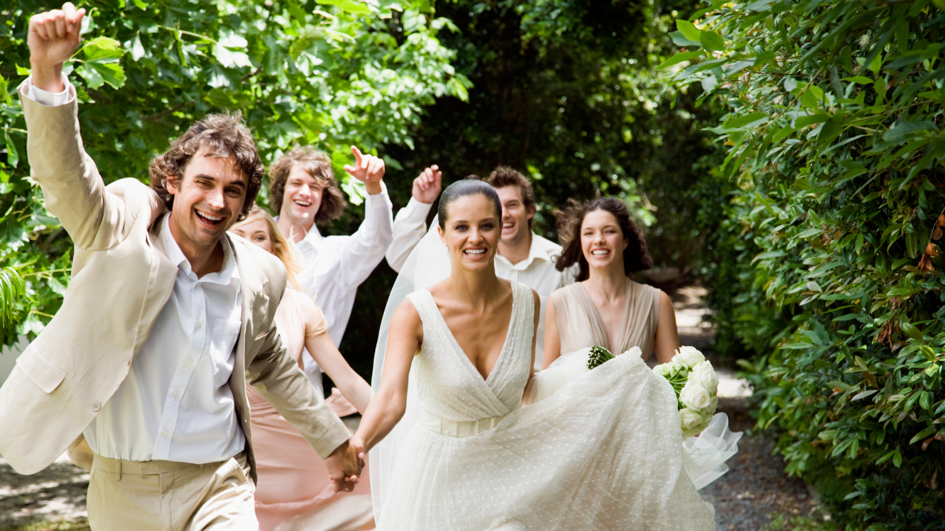 A joyful wedding party runs together along a lush garden path, with the bride and groom at the front.