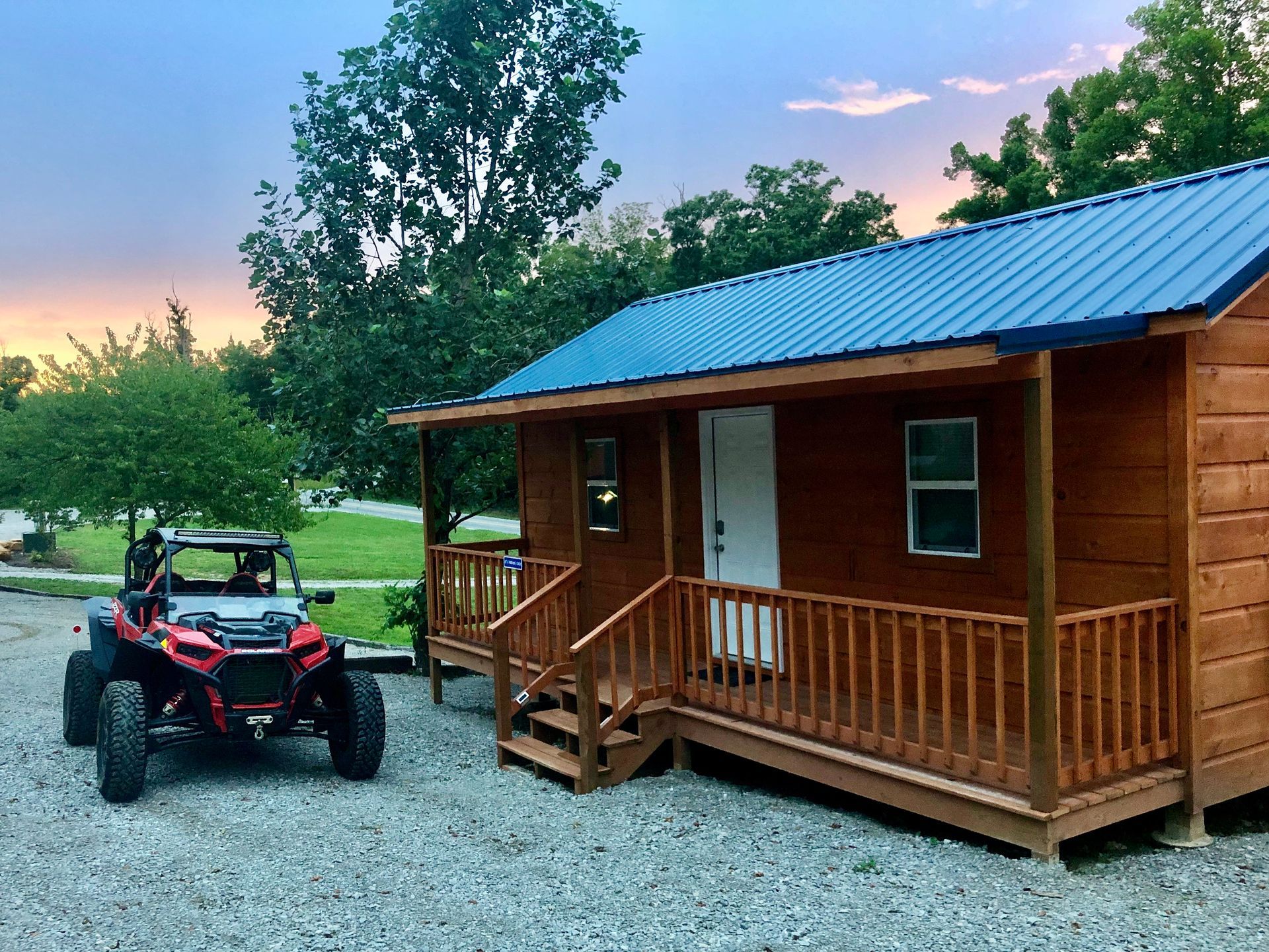 Small wooden cabin with blue metal roof beside a red-and-black off-road buggy at dusk