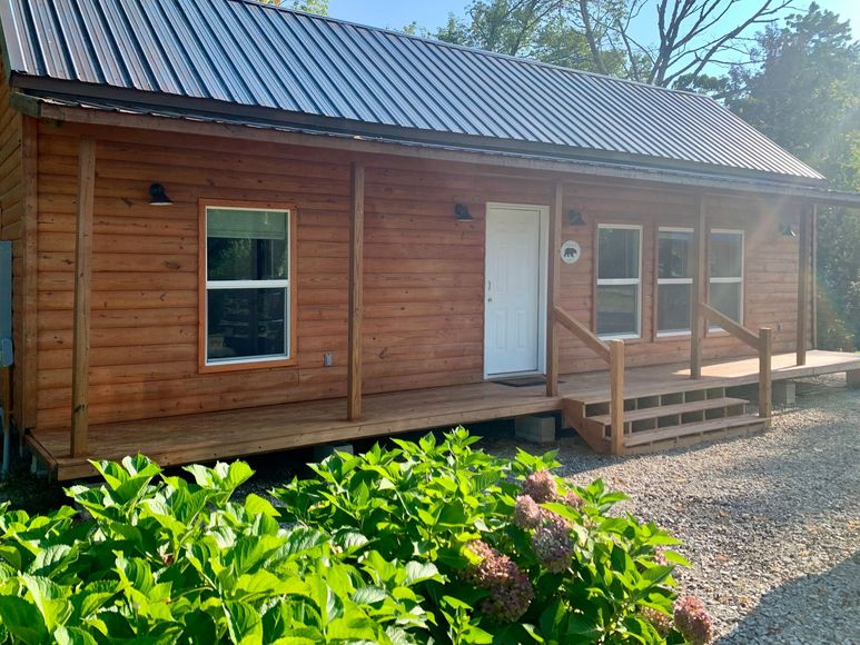 Wooden cabin with porch, white door, and gravel path, surrounded by green plants and flowers.