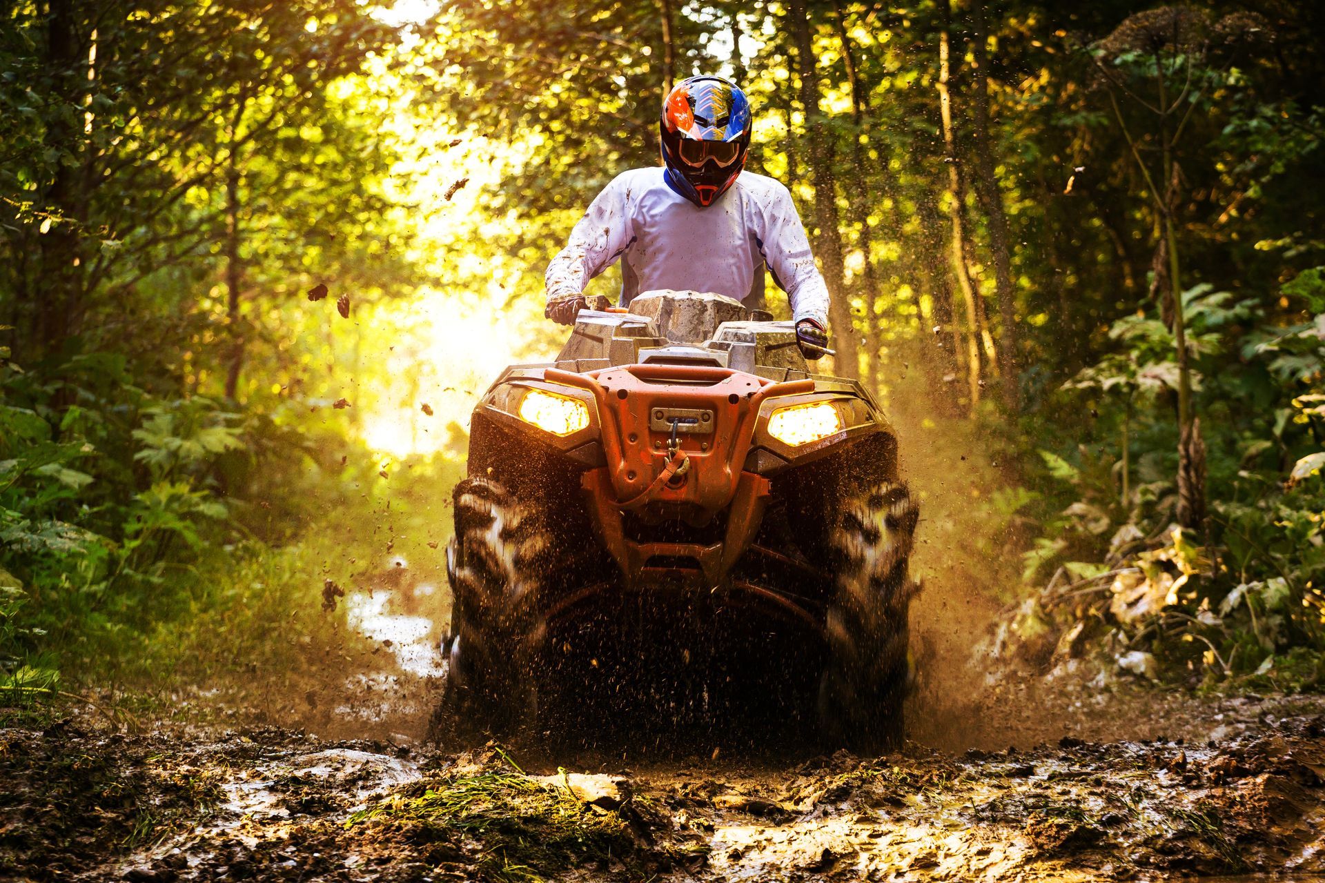 ATV rider driving through a muddy forest trail with sunlight streaming through trees