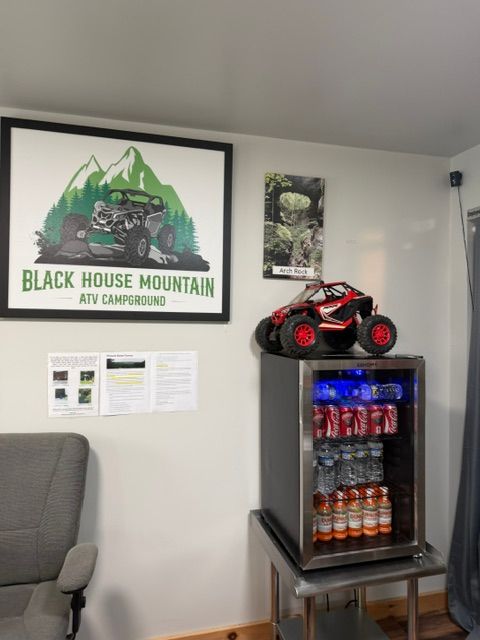 Office corner with Black House Mountain poster, mini fridge stocked with drinks, and a red toy ATV on top