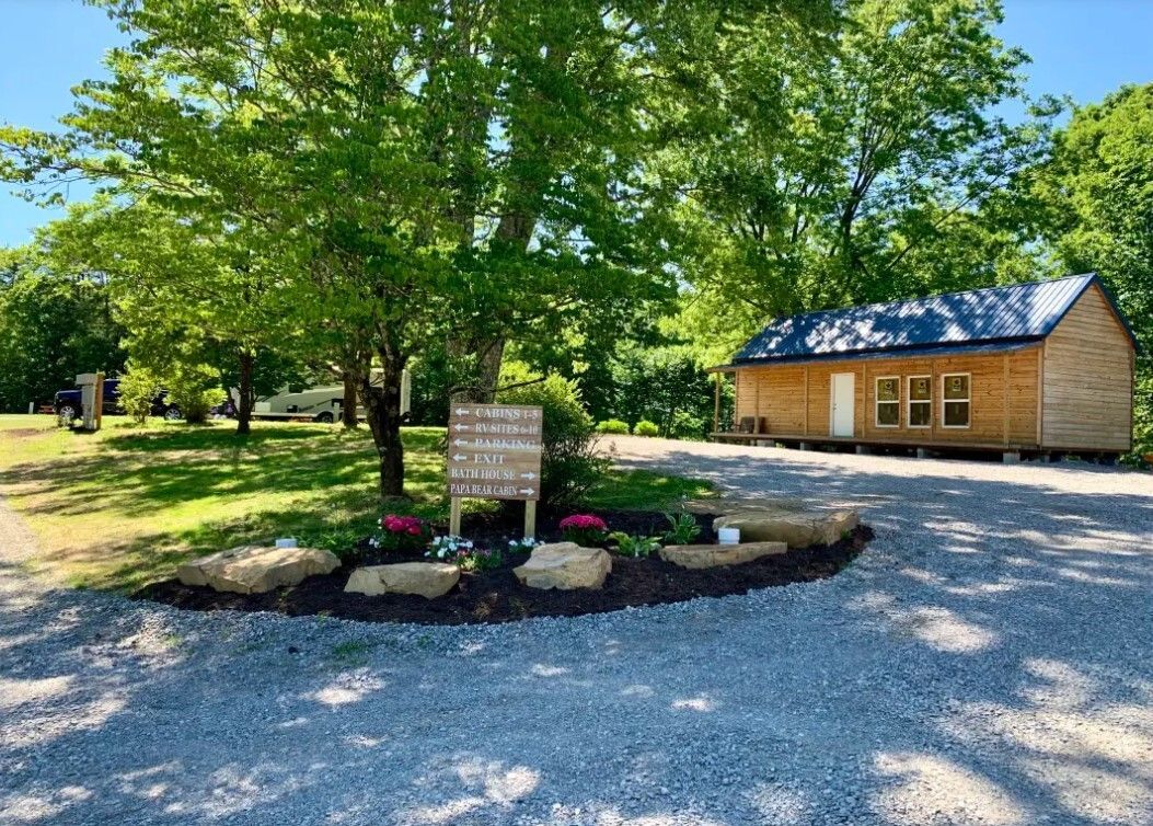Gravel driveway beside a small wooden cabin, with trees and a flowered stone border in front.