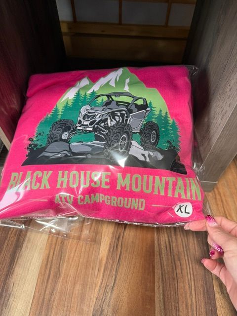 Pink T-shirt with an off-road vehicle graphic and “Black House Mountain Campground” text, held up indoors