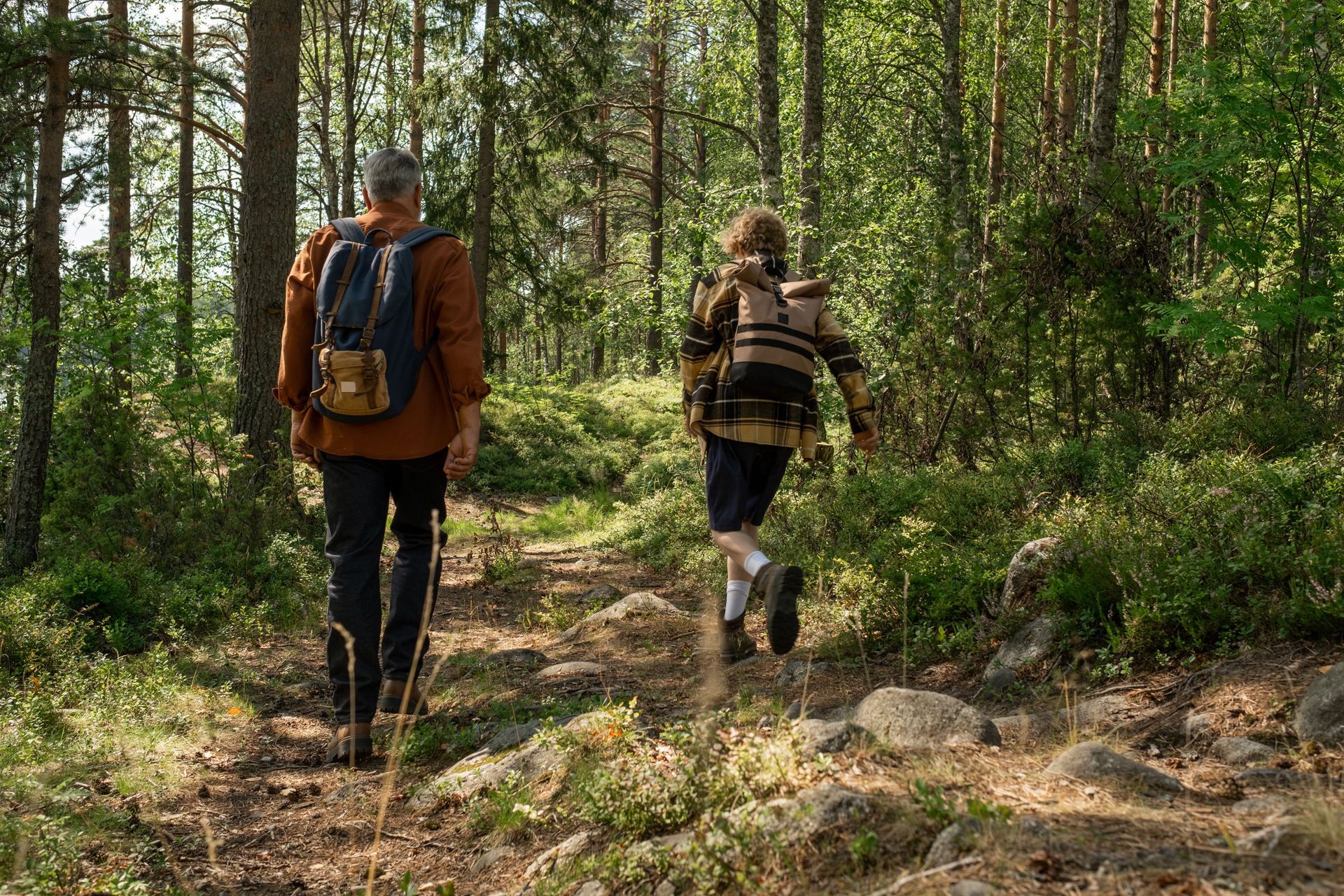 Two hikers with backpacks walking along a forest trail among tall trees and rocks