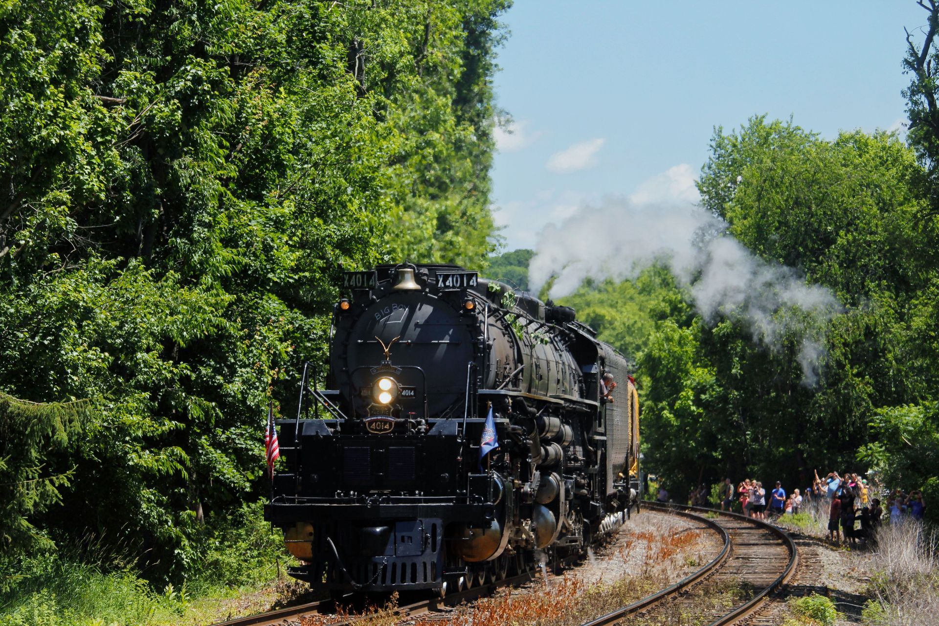 Steam locomotive rounding a curve on tracks through green trees, with smoke billowing behind.
