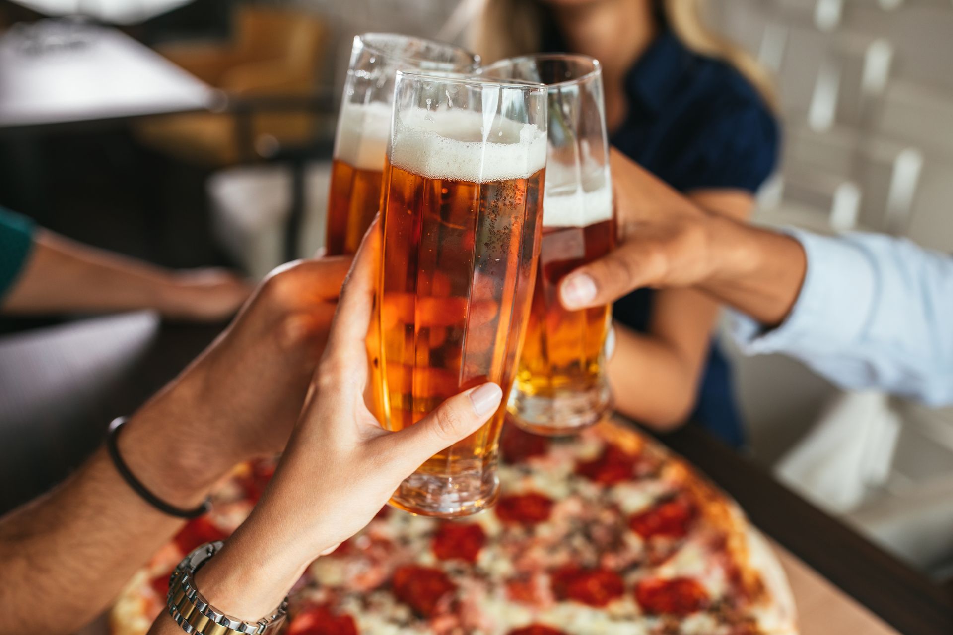 Hands clinking beer glasses over a pepperoni pizza at a restaurant table