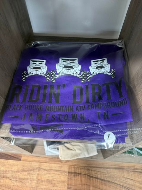 Purple DIRTY 30 race T-shirt in a box, with white logo graphics and folded clothes underneath