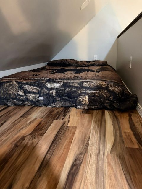 Small mattress with dark blanket in a slanted attic bedroom with wood-patterned floor and gray walls