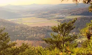 Sunlit valley with fields, forested hills, and mountains in the distance, seen through foreground trees