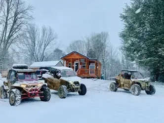 Three off-road buggies parked in snowy woods near a small wooden cabin.