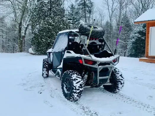 Black off-road buggy in snowy yard beside a wooden cabin, with snow-covered tires and roof rack.