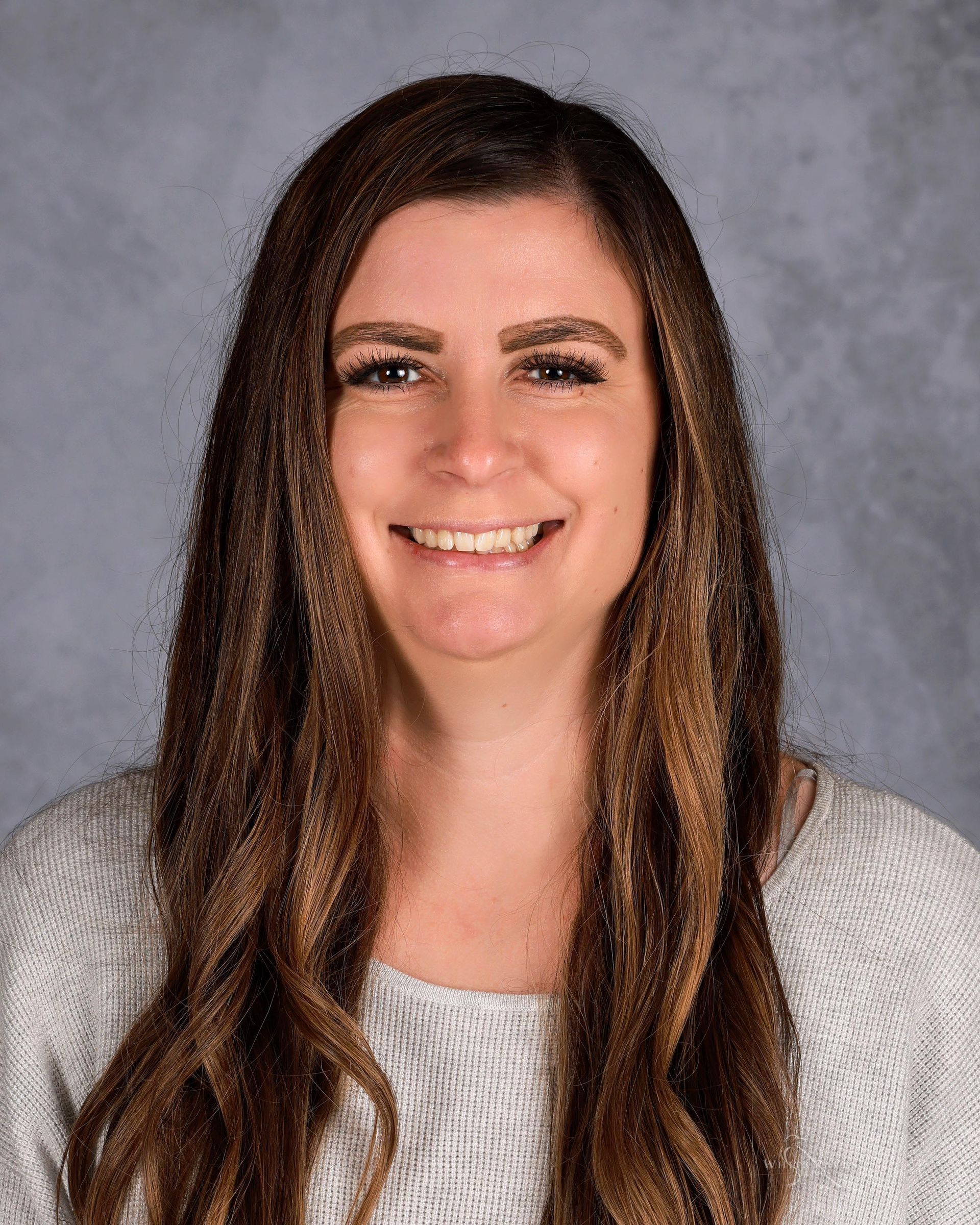 Woman with long brown hair smiling, wearing a gray sweater, against a blurred background.