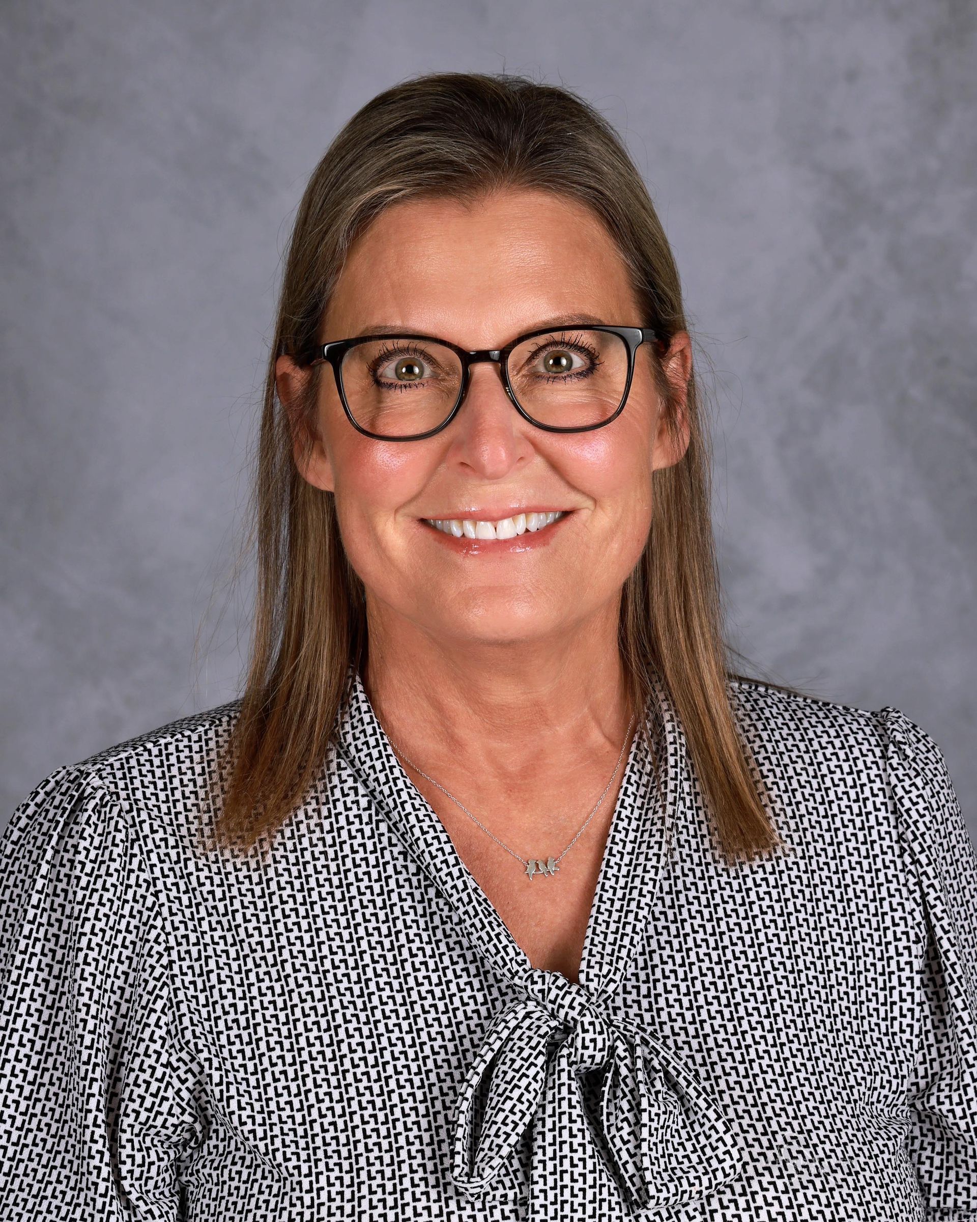 Woman with glasses smiles; wearing patterned blouse, against a grey backdrop.