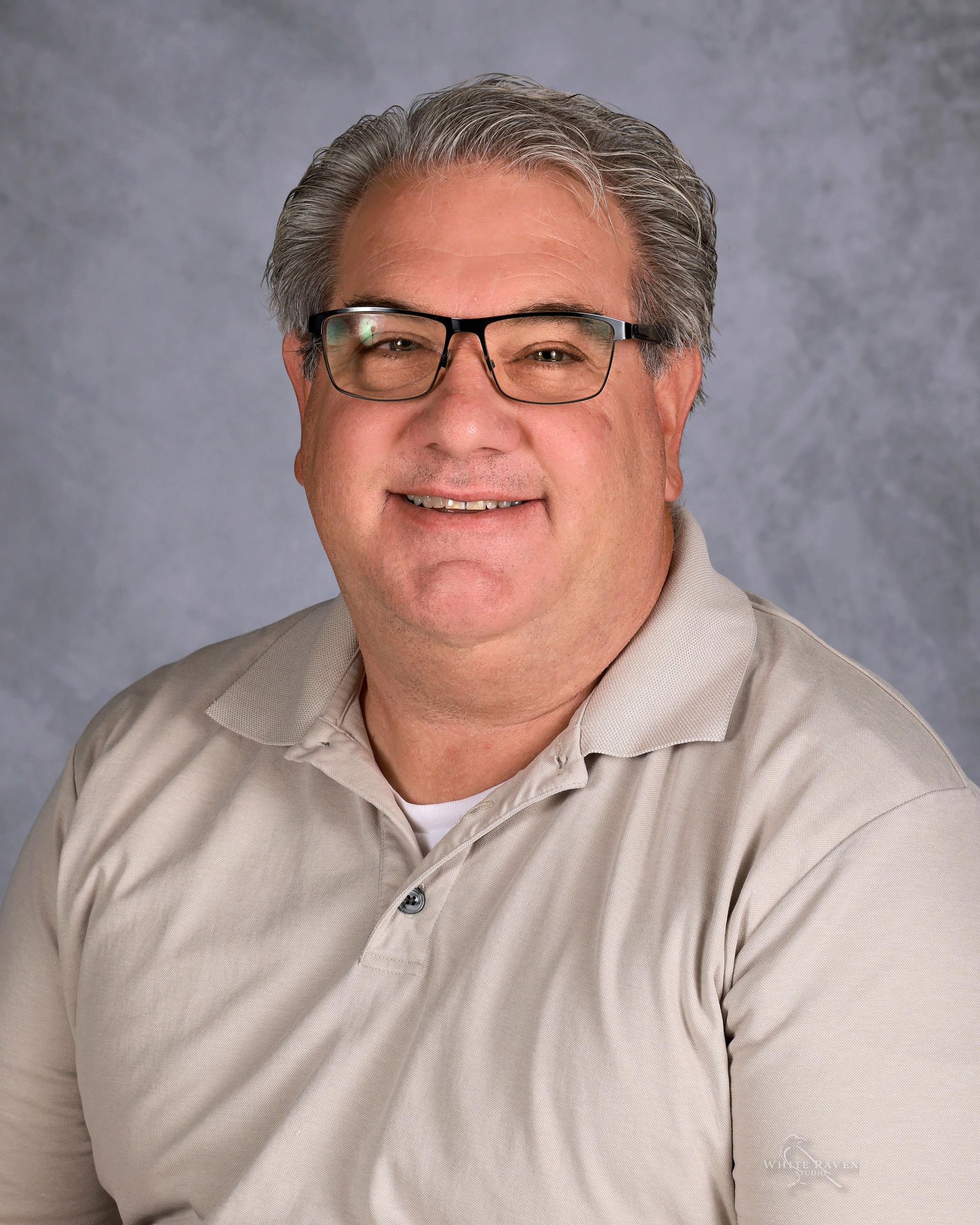 Man wearing glasses smiles, gray hair, light-colored collared shirt, against a soft-focus background.