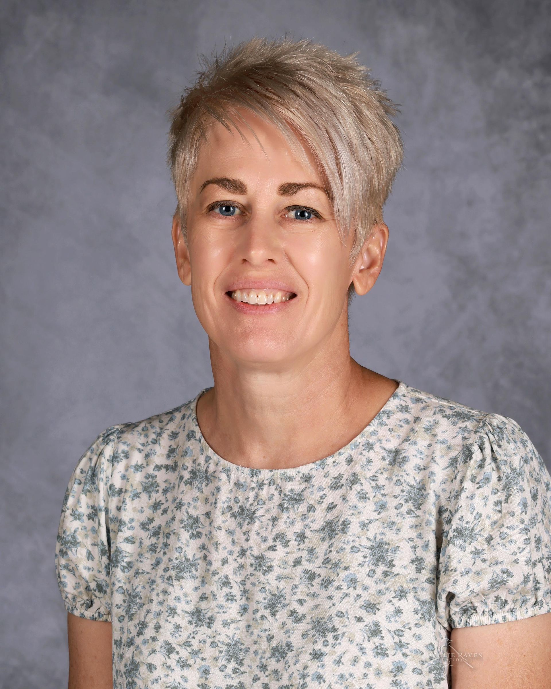 Woman with short, light-colored hair, smiling, wearing a floral print top, against a gray background.