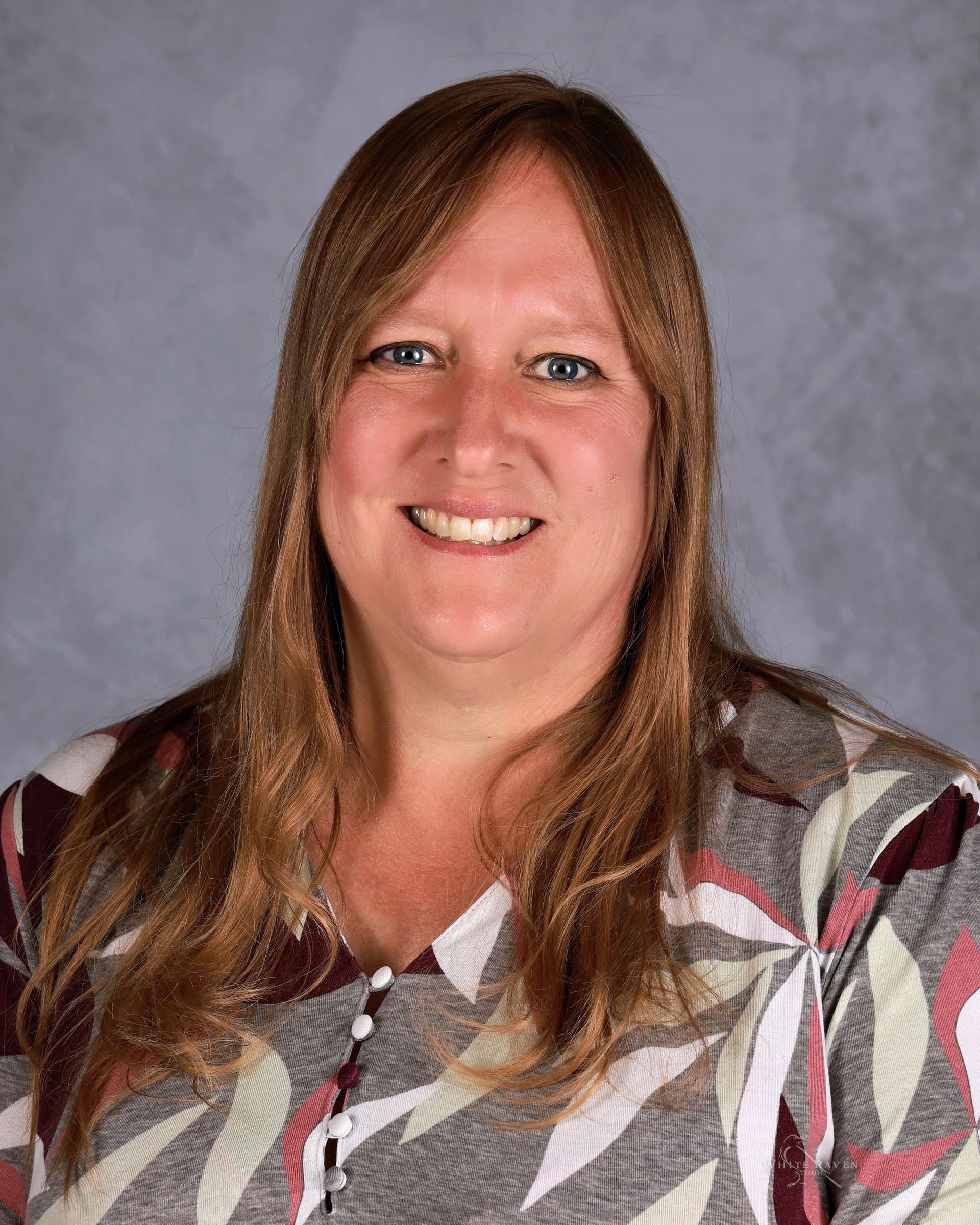 Woman with long brown hair, smiling, wearing a patterned blouse, against a gray background.