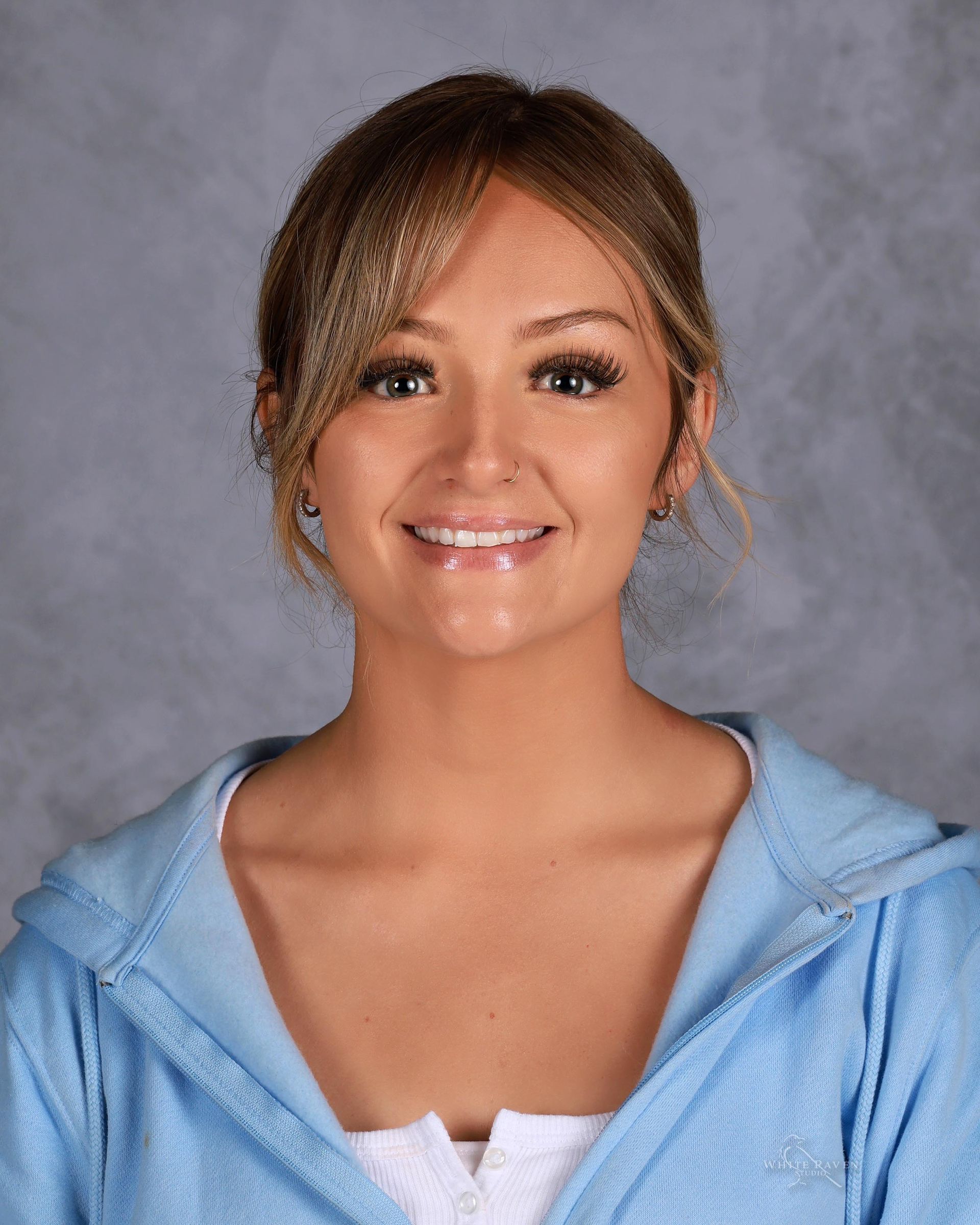 Woman smiling, wearing a light blue hooded sweatshirt against a gray background.