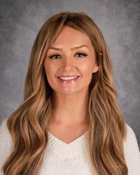Smiling woman with long blonde hair against a gray studio backdrop, wearing a white top