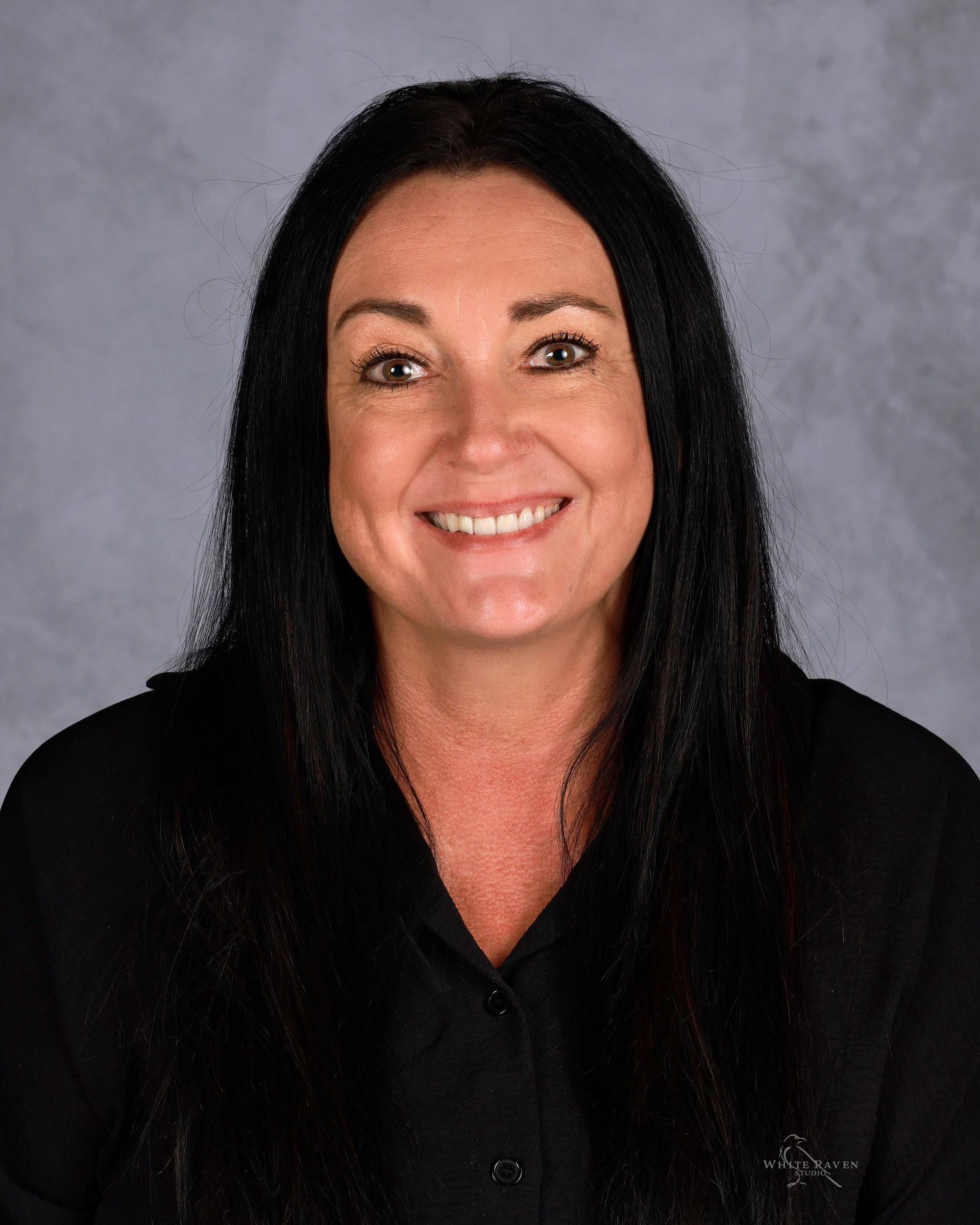 Woman with dark hair smiling, wearing a black shirt, in front of a gray backdrop.