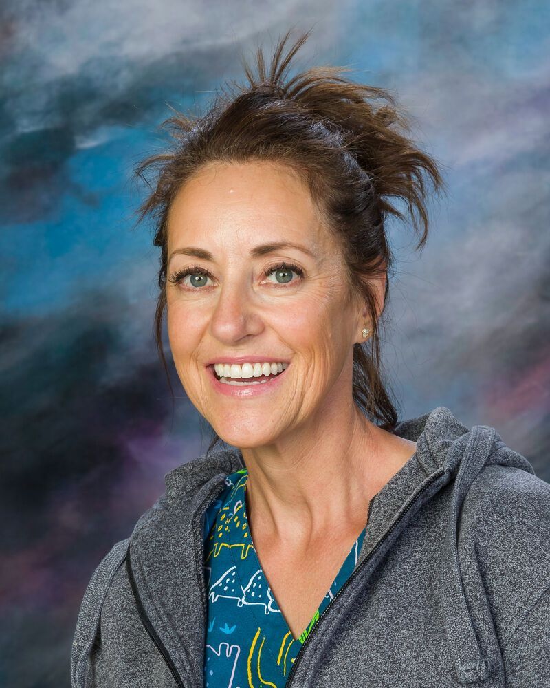 Woman smiles broadly, wearing a gray hoodie over a teal shirt with a pattern. Background is a blurred blue and gray.
