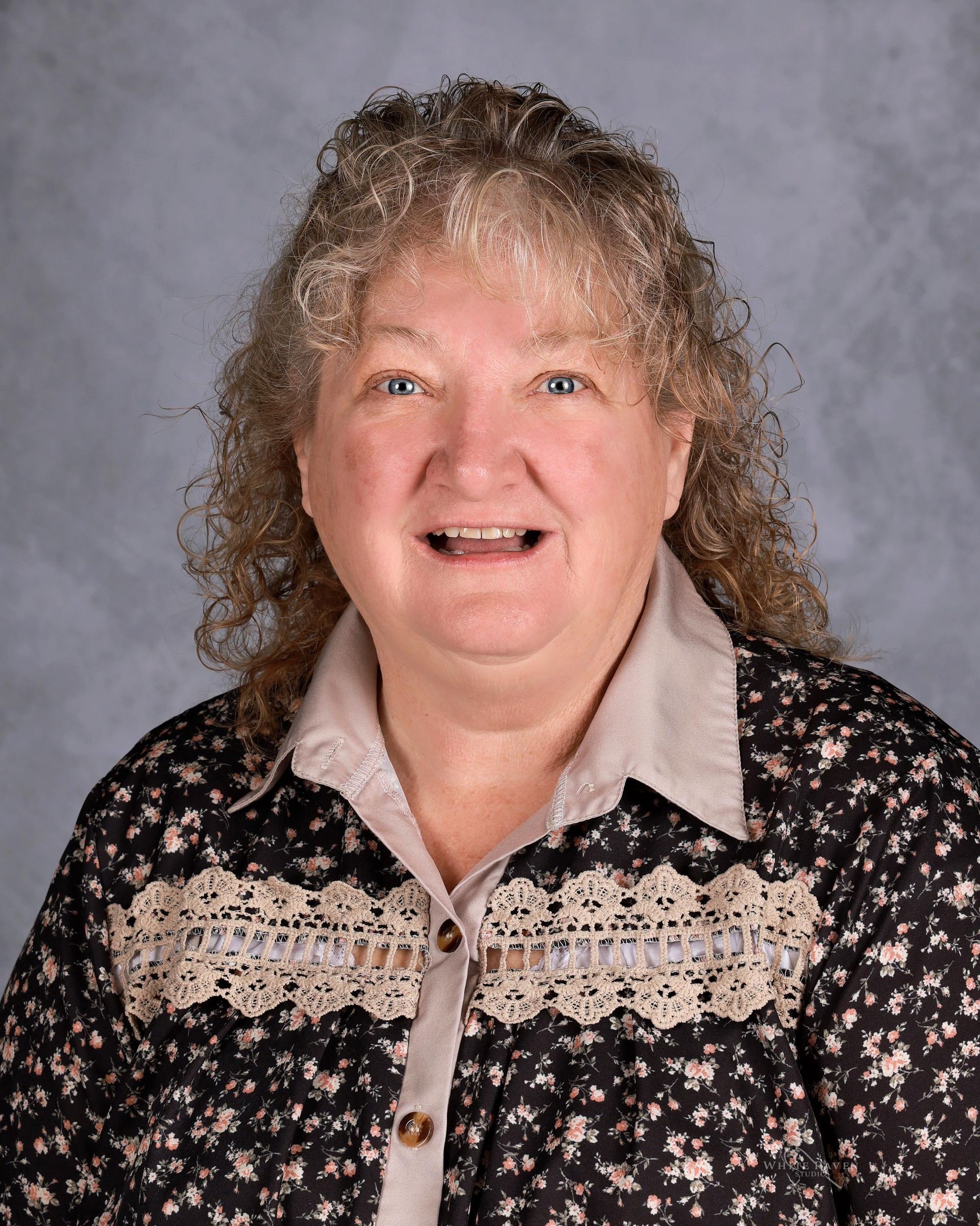 Woman with curly hair smiles, wearing floral shirt with beige collar, against a gray background.