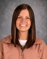 Portrait of a smiling person with straight brown hair, wearing a tan jacket against a gray backdrop