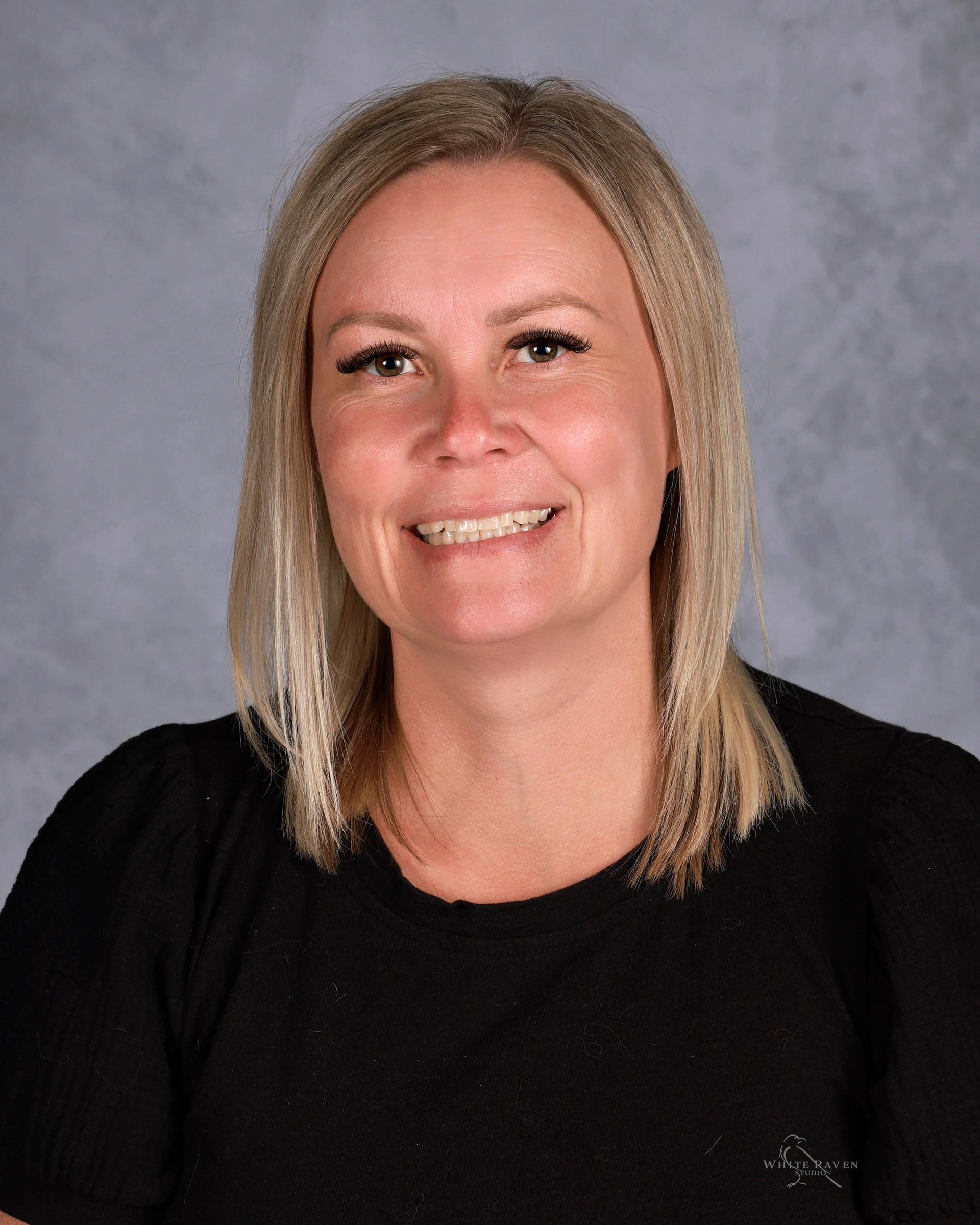 Blonde woman smiling, wearing a black shirt, against a blurred gray background.