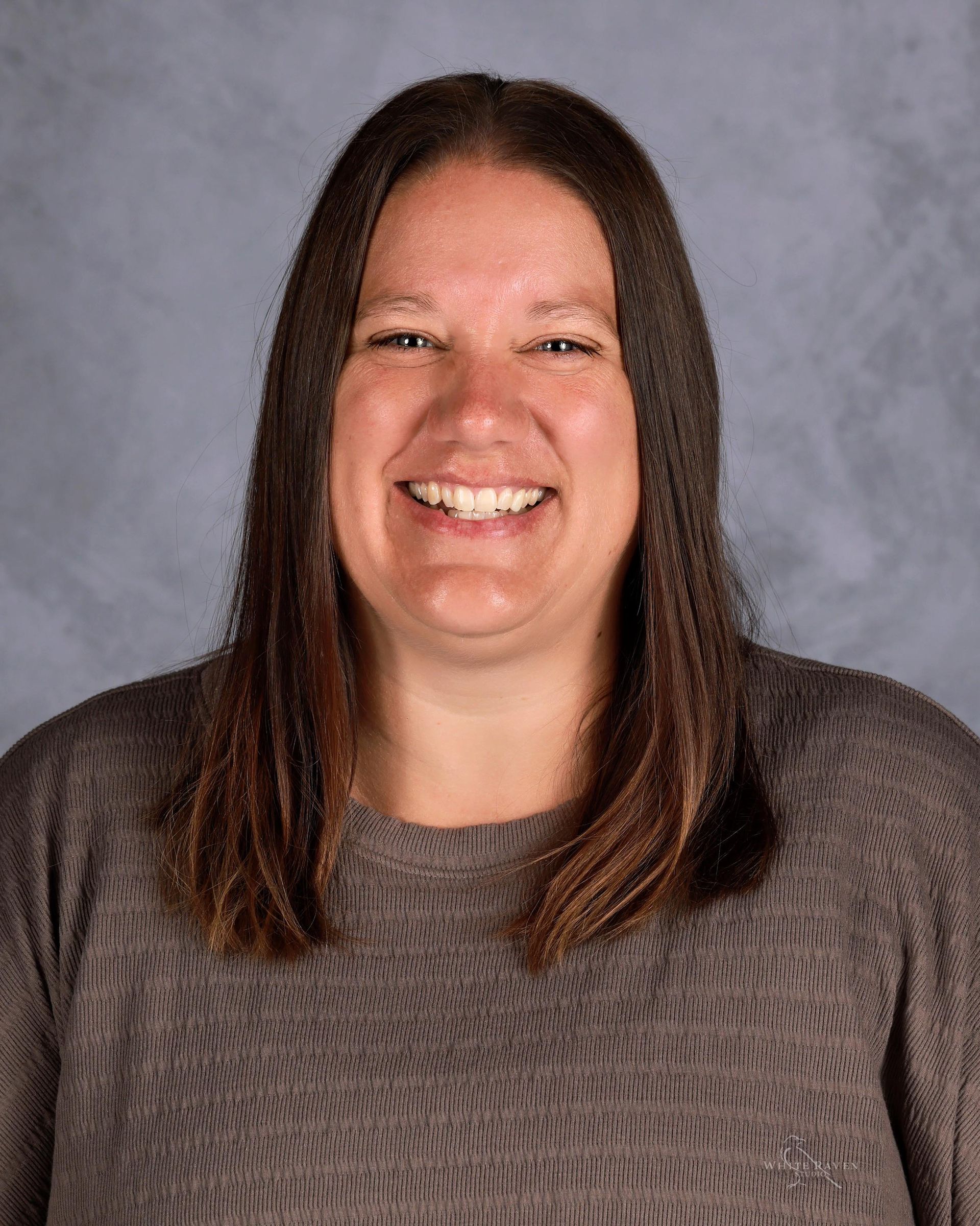 Woman smiling at the camera, wearing a gray sweater. Brown hair against a gray backdrop.