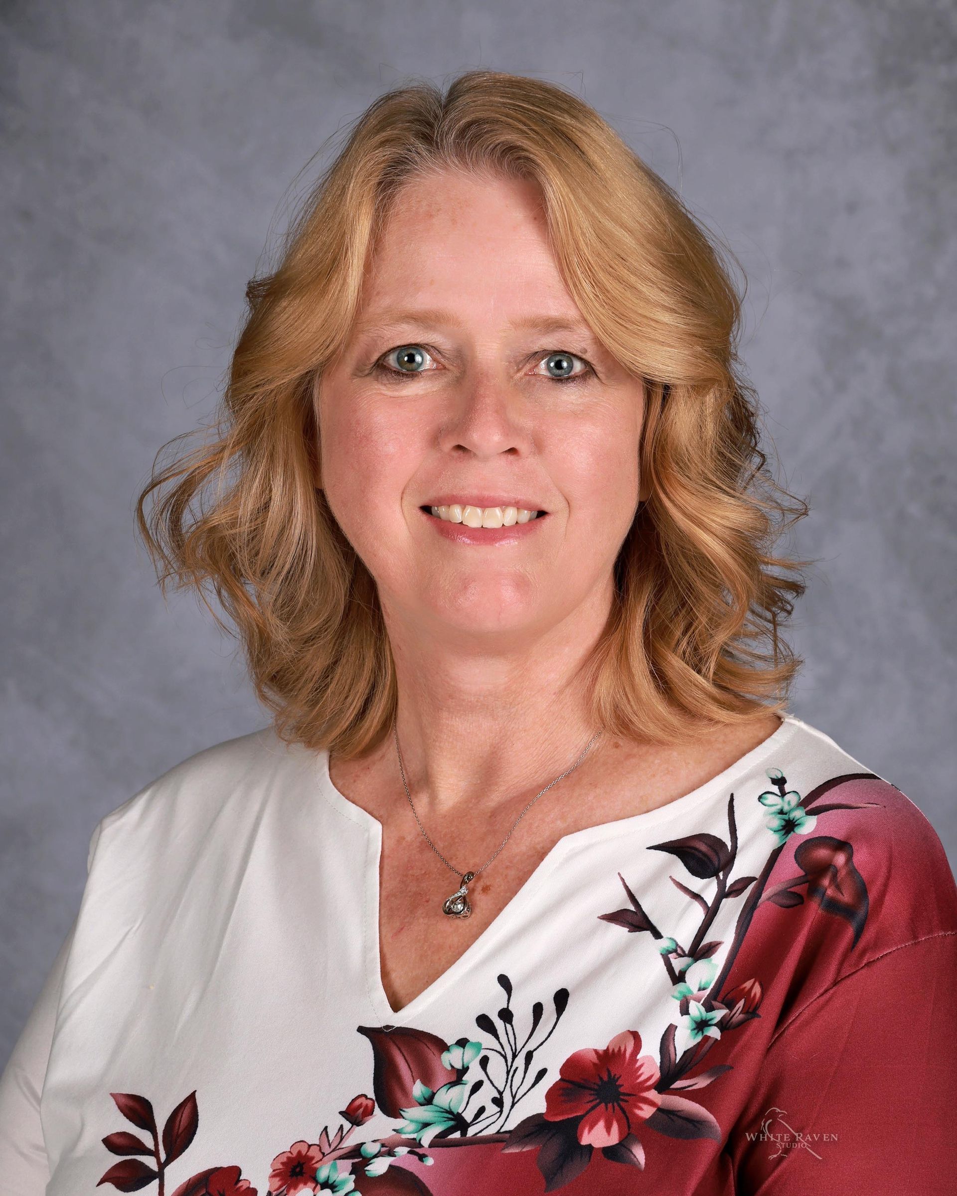 Woman with blonde hair wearing a white and burgundy floral top smiles at the camera.