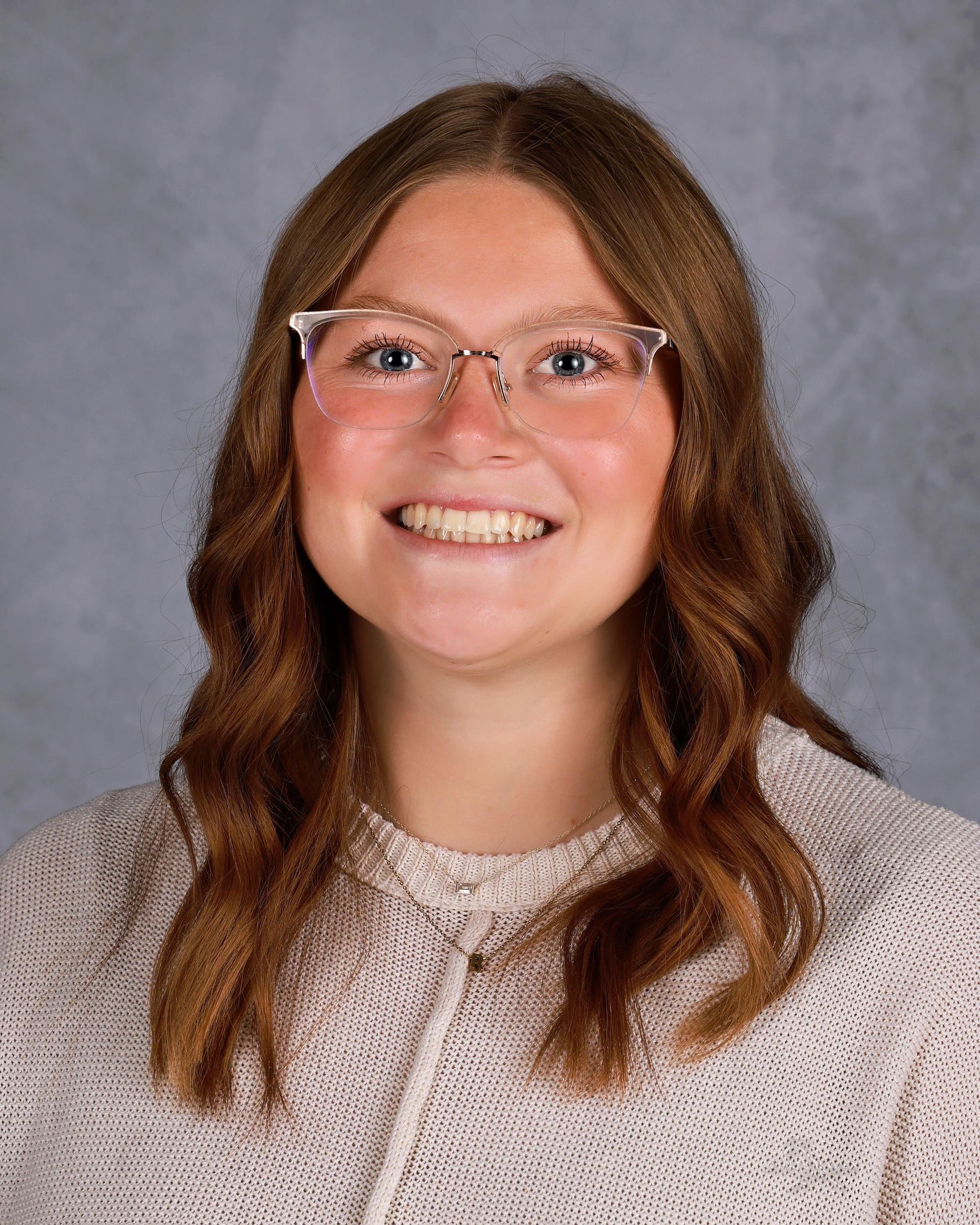 Woman with light brown wavy hair wearing glasses, smiling at the camera, light-colored sweater, grey background.