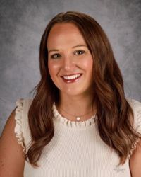 Smiling person with long wavy brown hair in a cream blouse against a gray studio backdrop