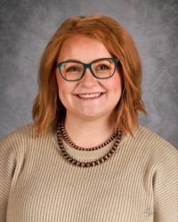 Smiling person with red hair and glasses wearing a beige sweater against a gray backdrop