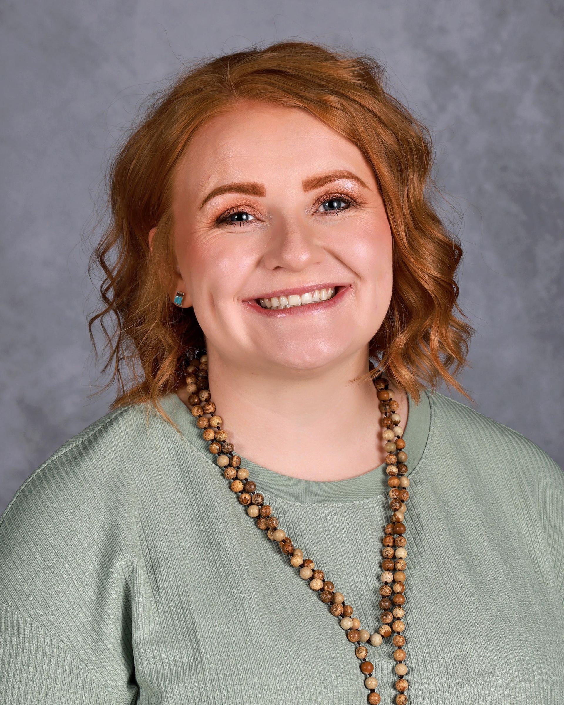 Woman with red hair smiling, wearing a green sweater and wooden bead necklace. Gray background.