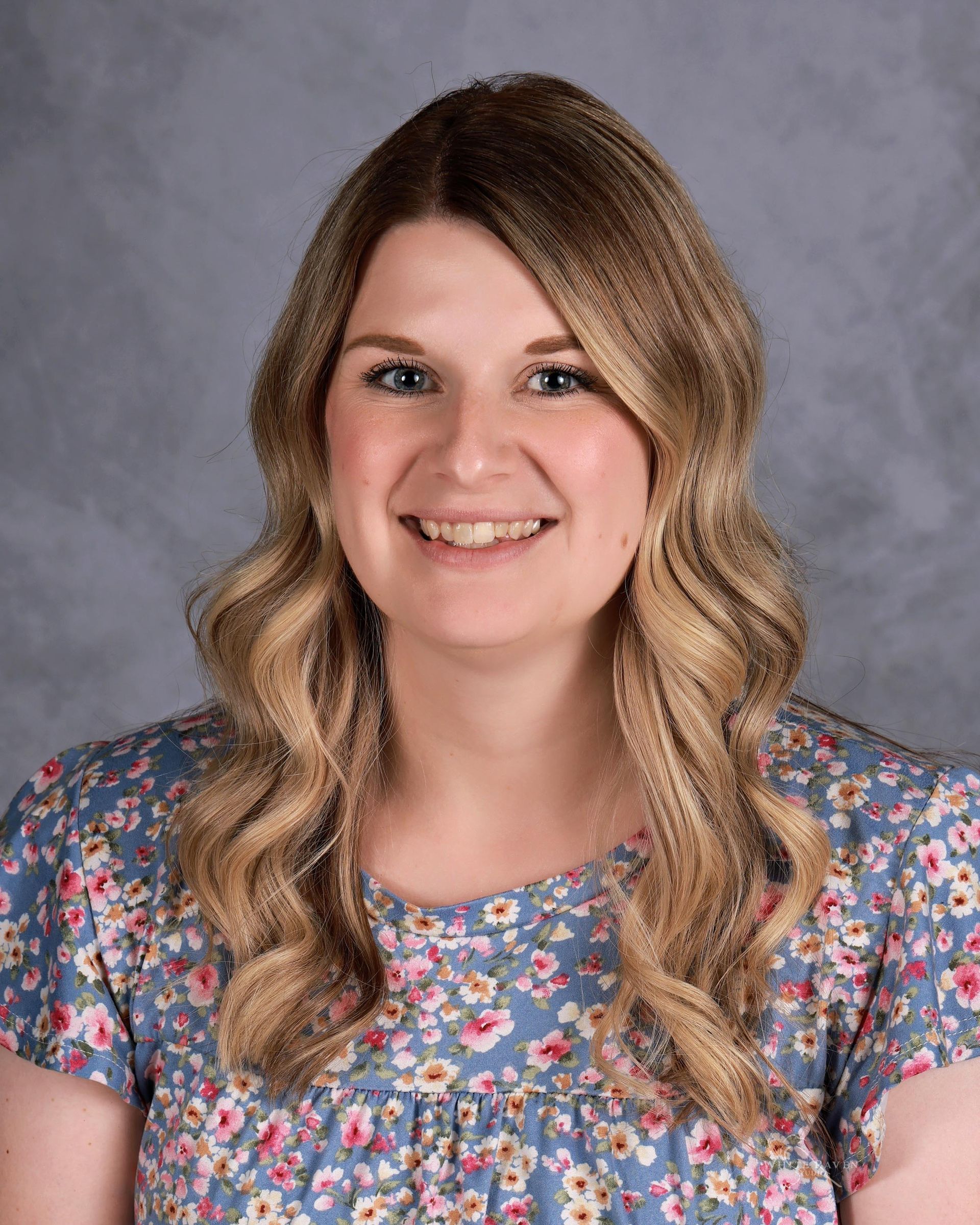 Woman with wavy blonde hair smiling, wearing a blue floral top, against a gray background.