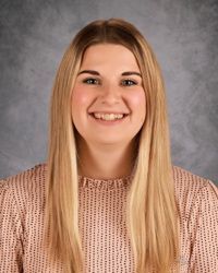 Smiling person with long blonde hair in a pink patterned top against a gray studio background