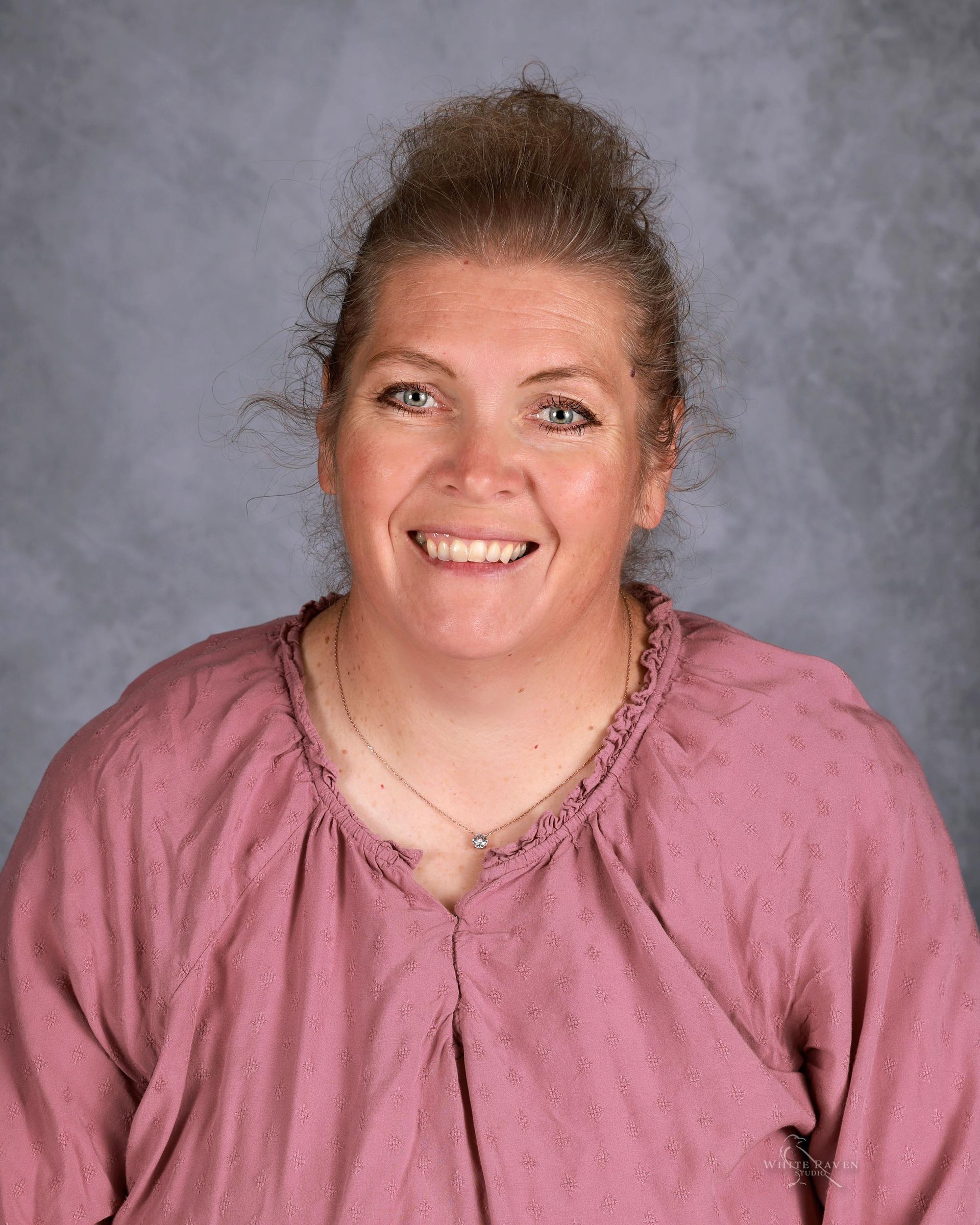 Woman with light skin, smiling, wearing a pink top, hair pulled back, against a grey background.