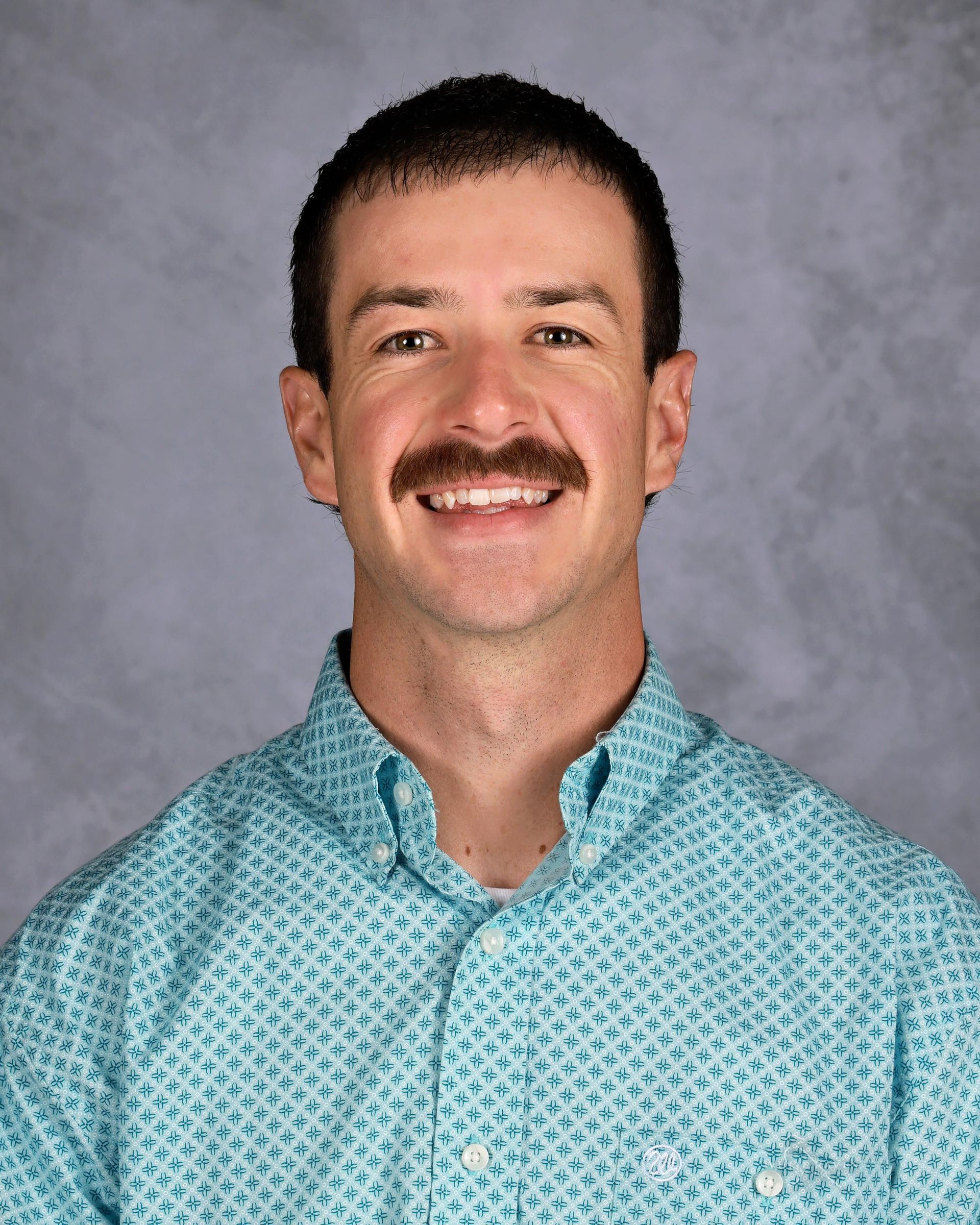 Man with a mustache smiles, wearing a turquoise patterned button-down shirt.