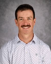 Headshot of a smiling man with a mustache wearing a white patterned shirt against a gray backdrop