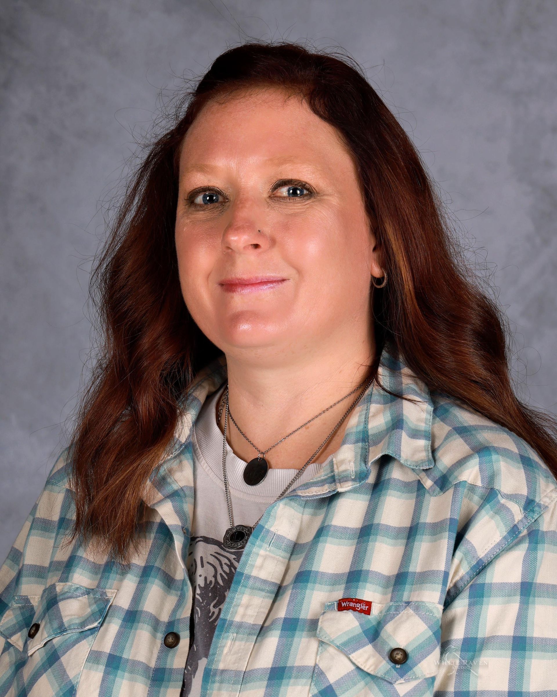 Woman with reddish-brown hair, wearing a plaid shirt, smiling, posing in front of a gray background.