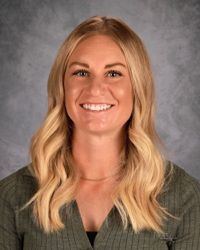 Studio headshot of a smiling blonde woman against a gray backdrop, wearing a green top.