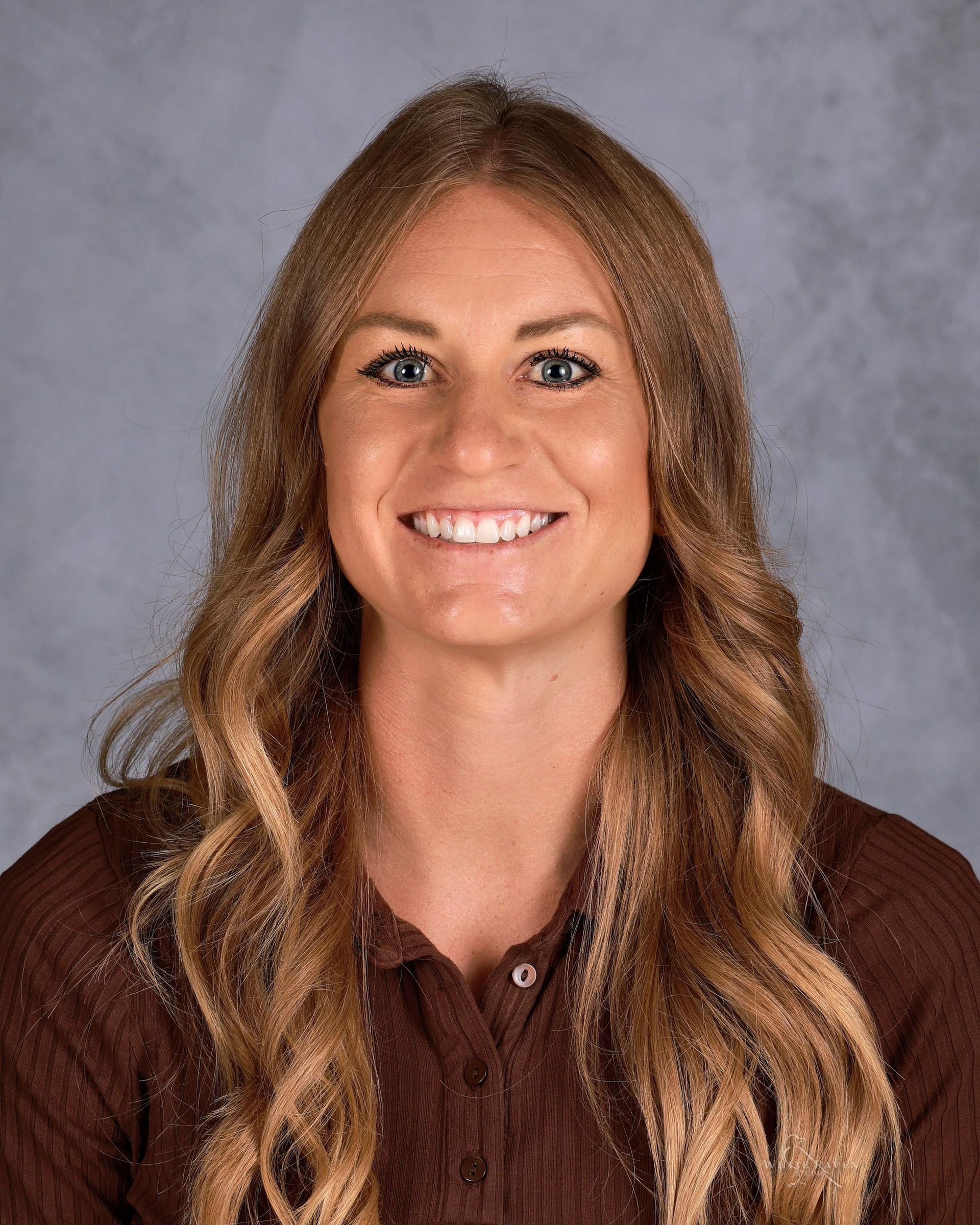 Woman with long blonde hair, smiling, wearing a brown shirt against a gradient background.