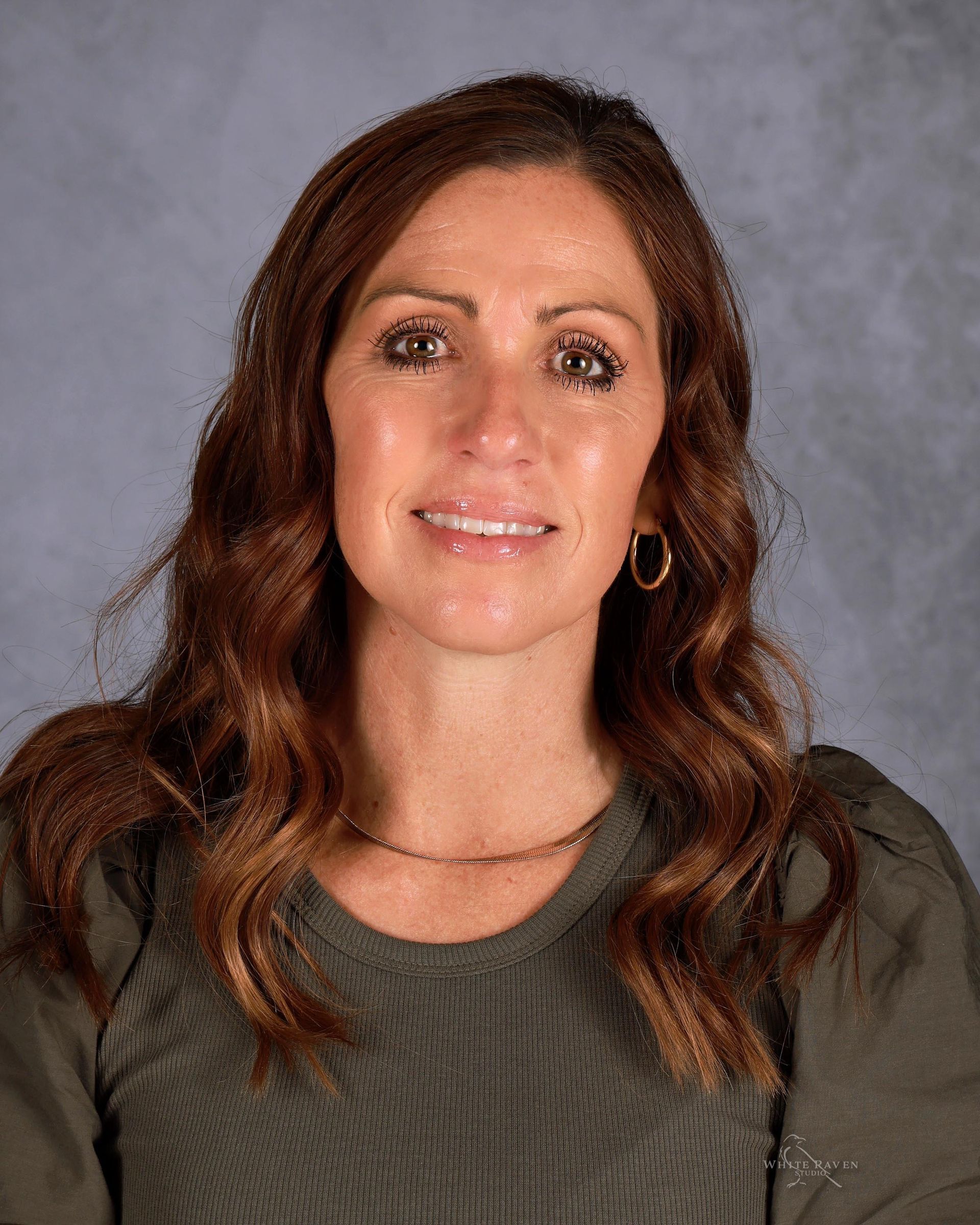 Woman with long, wavy brown hair smiles. She wears a green sweater and a small necklace, set against a blurred gray background.