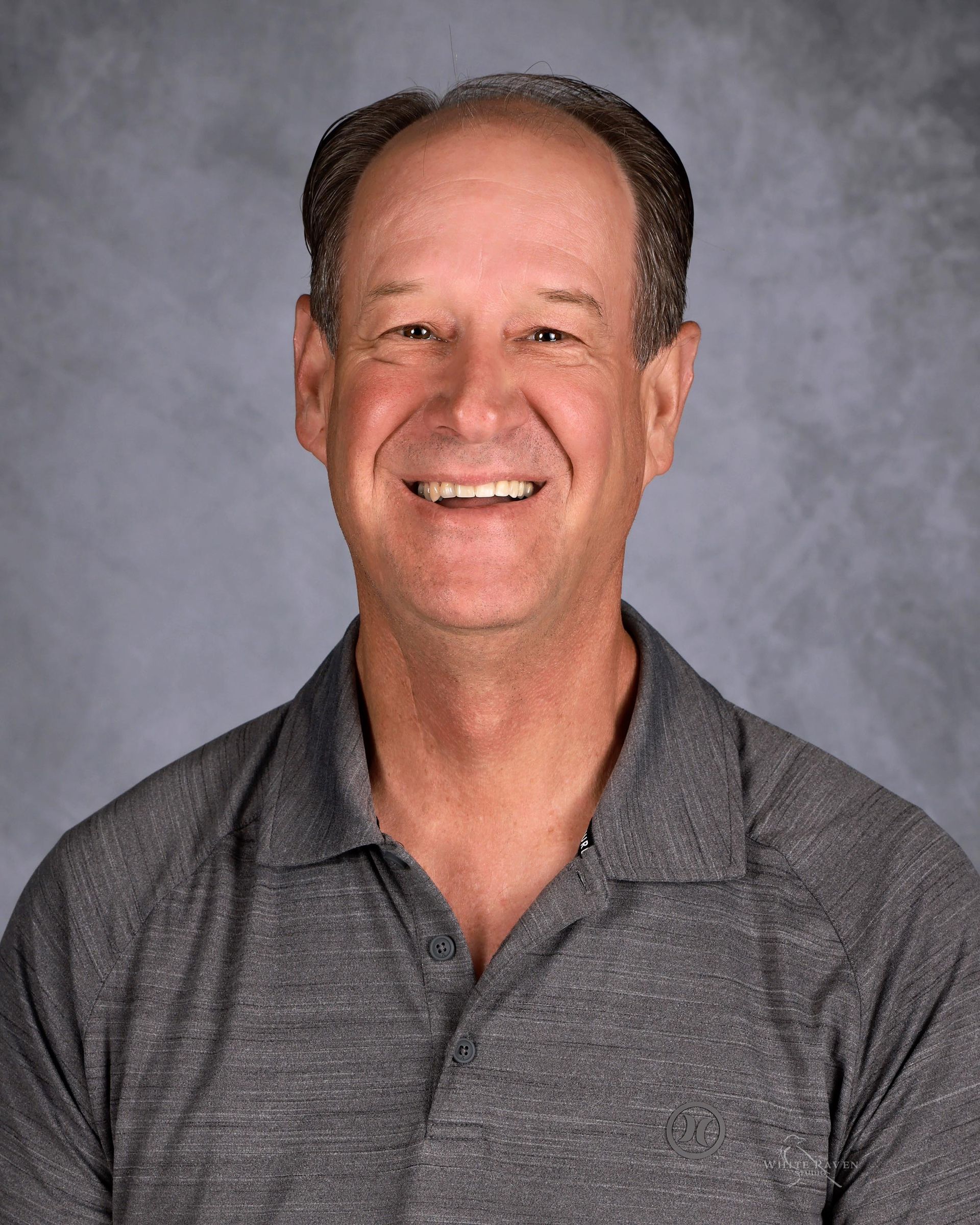 Man smiling, wearing a gray collared shirt. Against a blurred, neutral background.