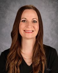 Professional headshot of a smiling person with long brown hair on a gray studio background