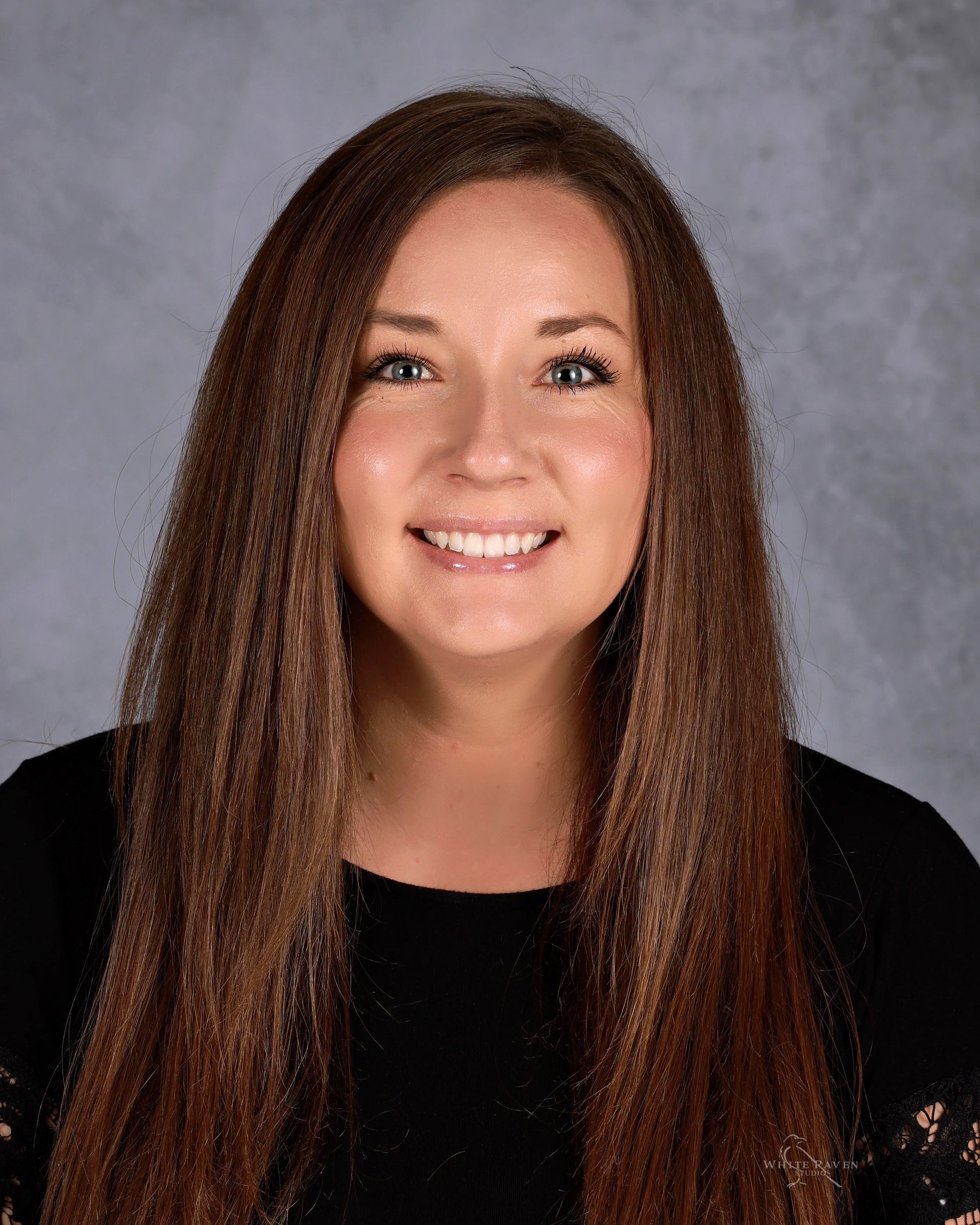 Woman with long brown hair smiles at the camera, wearing a black top. Background is a gradient gray.